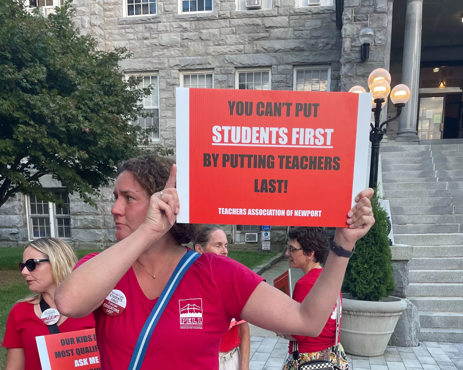 A Teachers Association of Newport (TAN) member wears a serious expression and a red shirt as they stand on a picket line with a sign reading "You can't put students first by putting teachers last!"