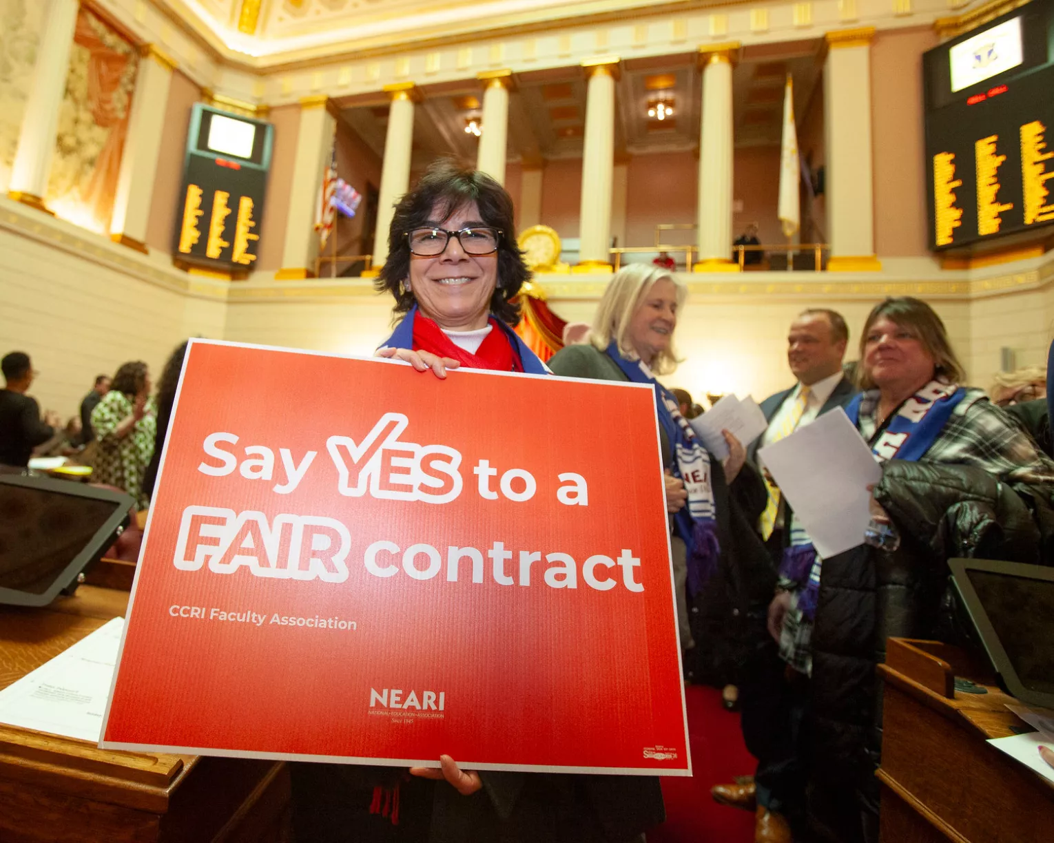 A NEARI CCRI member, Professor Jeanne Mullaney, on the floor at the Statehouse with a sign that says "Say Yes to a FAIR contract."