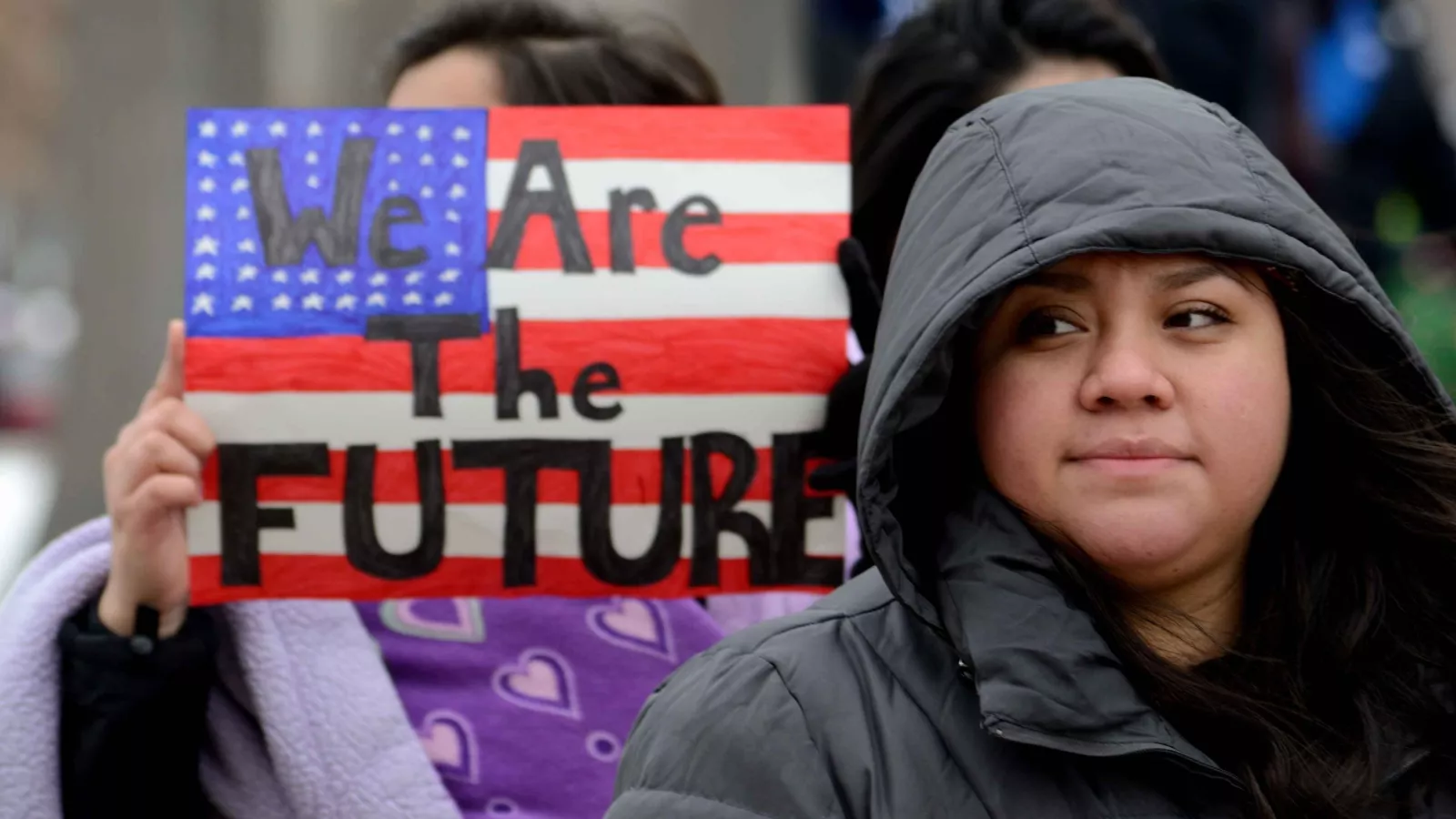 A student in a black puffy coat standing in front of a small poster that looks like the American flag. The poster says "We Are the Future."