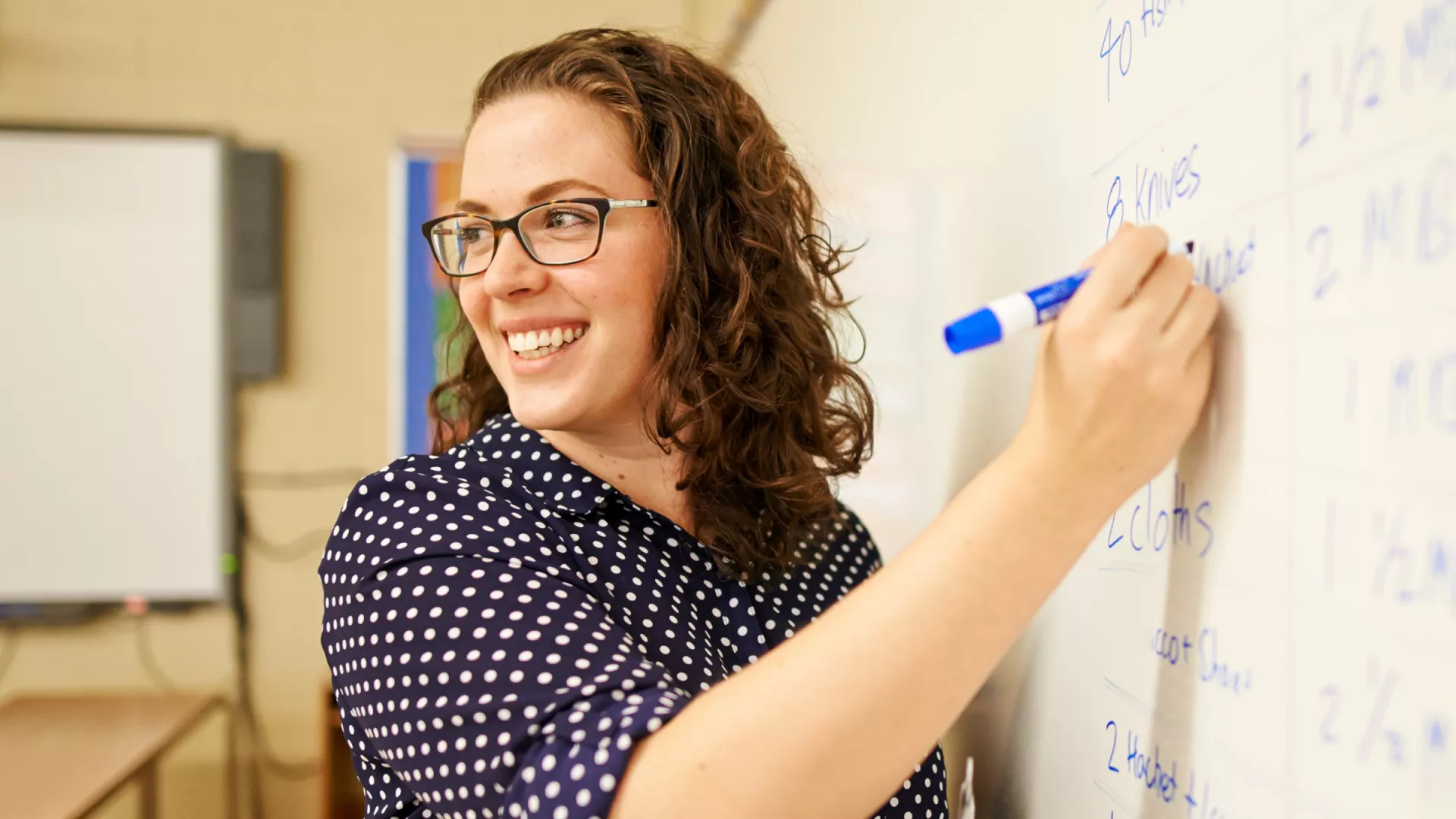 An educator writes on the large dry erase board at the front of a classroom.