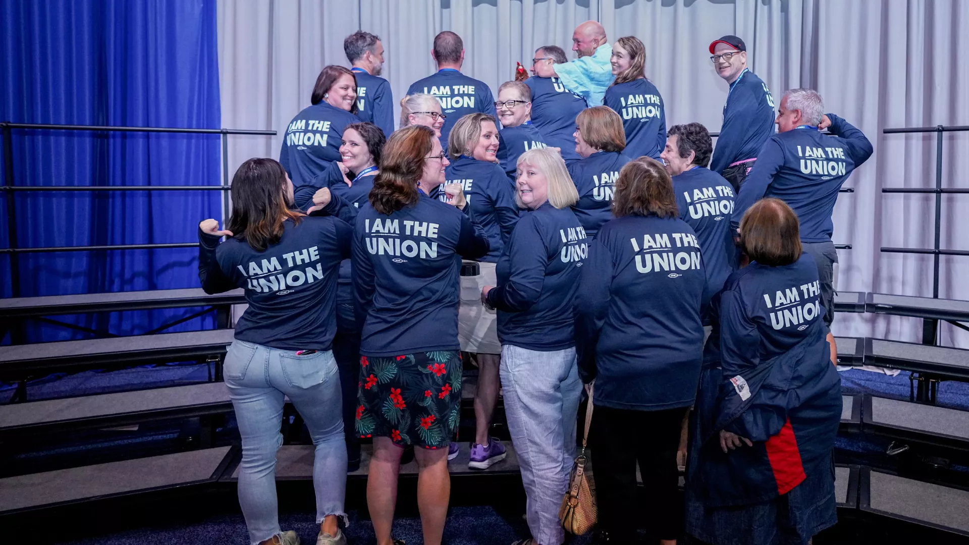 NEARI 2023 NEA RA delegates pose for a delegation picture with their backs to the camera. They are all pointing to the words on the back of their navy shirts, which say, "I am the union!" in white block text. You can see some people smiling and laughing in profile as they look over their shoulders.