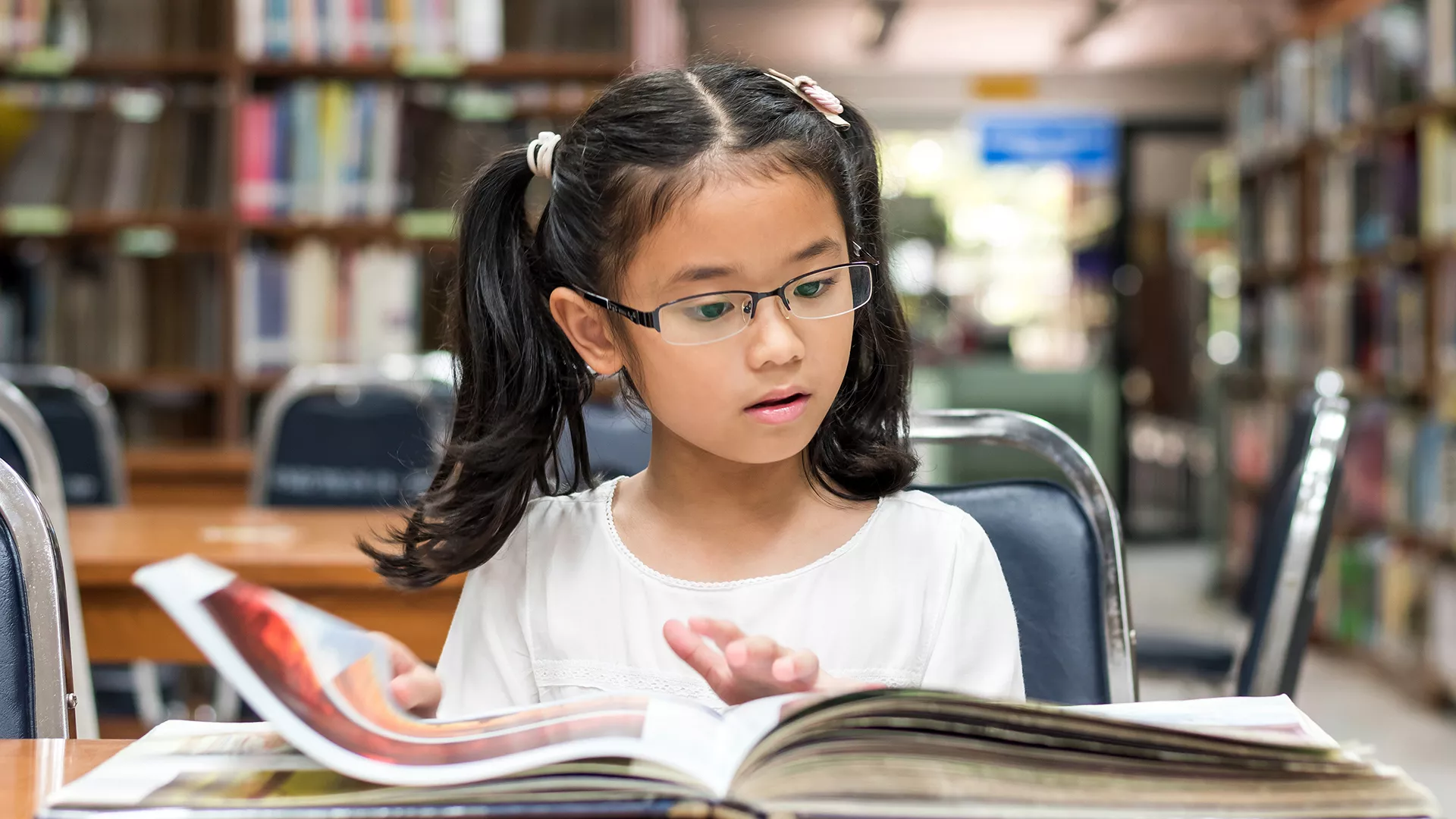 young girl in glasses sits and reads a picture book at library