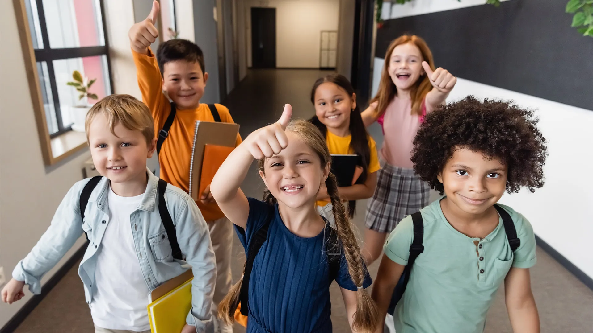 6 elementary students walk down a school hallway with thumbs up looking at the camera