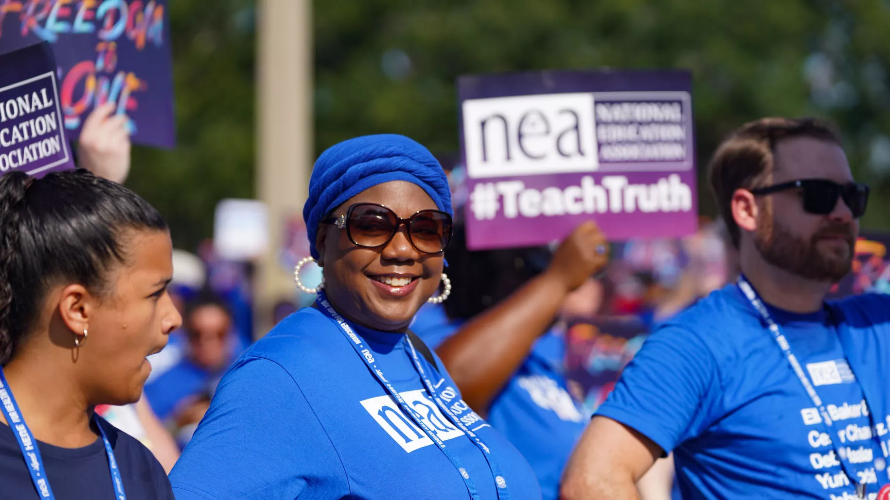 A Black woman in an NEA t-shirt smiles at the camera. She surrounded by people holding signs that say "Freedom to Love" and "teach Truth"