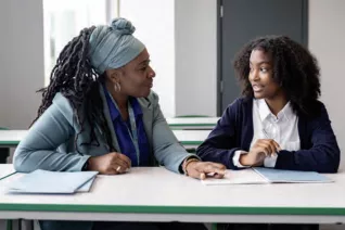 Black female teacher and student sit at a desk and discuss schoolwork