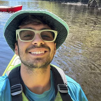 A man wearing a hat and sunglasses smiles as he takes a selfie outdoors.