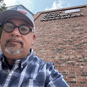 A man with glasses and a hat looks directly to camera while taking a selfie