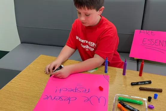 A child wearing a red shirt sits at a small wood table working intently on making a sign that says "We deserve respect!" using markers on a pink paper.