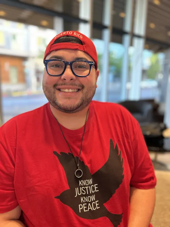 A man wearing a red shirt adorned with a bird graphic and the message "Know Justice Know Peace"smiles.
