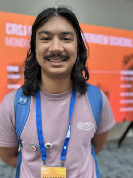 A man with long hair wearing a light purple shirt and a blue backpack stands smiling in a convention setting.