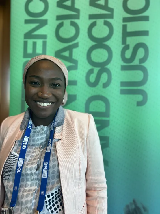 A woman in a light-biege jacket stands in front of a green banner, smiling confidently.