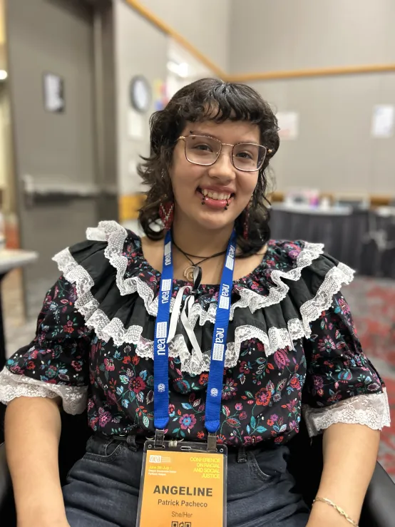 A woman with glasses sits in a chair, wearing a colorful floral shirt, smiling softly at the camera.