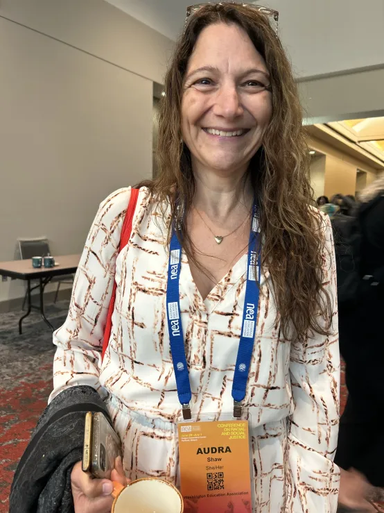 A woman wearing a badge holds a cup of coffee, smiling as she stands in a conference setting.