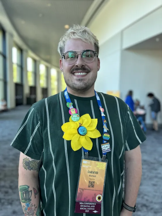 A man wearing a yellow flower necklace smiles in a convention setting.