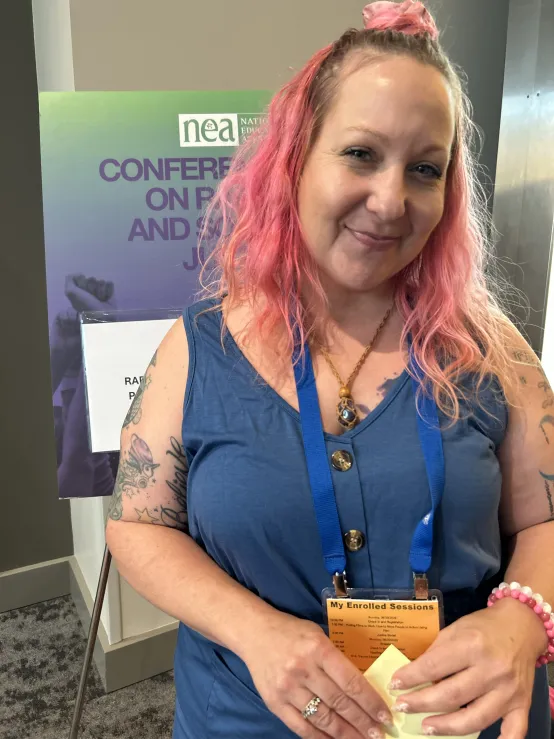 A woman with pink hair is holding a piece of paper, smiling at the camera..