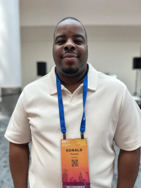 A man wearing a blue lanyard displaying a name tag around his neck, standing in a conference setting.