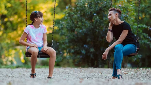 A mother and daughter sit on swings, looking at each other as they talk.