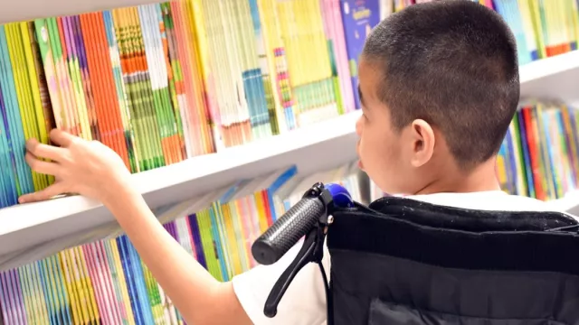 A boy in a wheelchair looks at brightly colored books on a library shelf.