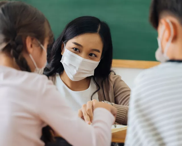 Asian teacher in mask working with 2 students in classroom