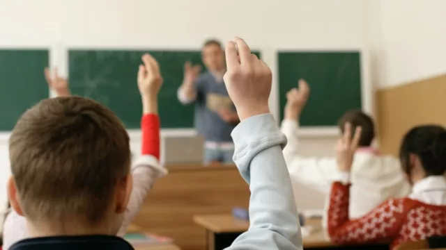 Students are raising hands in a classroom. The picture is taken from the back of the classroom, so you can see the backs of the children and their hands. The teacher up front at the chalkboard is out of focus.